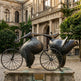 Bronze sculpture of two figures on a bicycle in front of an ornate building.