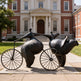Bronze sculpture of two figures on a bicycle in front of a brick building
