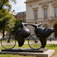 Sculpture of two figures on a bicycle in front of an architectural building