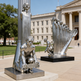 Metallic sculpture of a person with a ball and another with a harp-like instrument in front of a building.
Reflective metallic artwork showing multiple human forms emerging upward from a sculpted hand