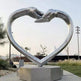 Symbolic metal heart artwork with raised hands touching at the top, set on a concrete base