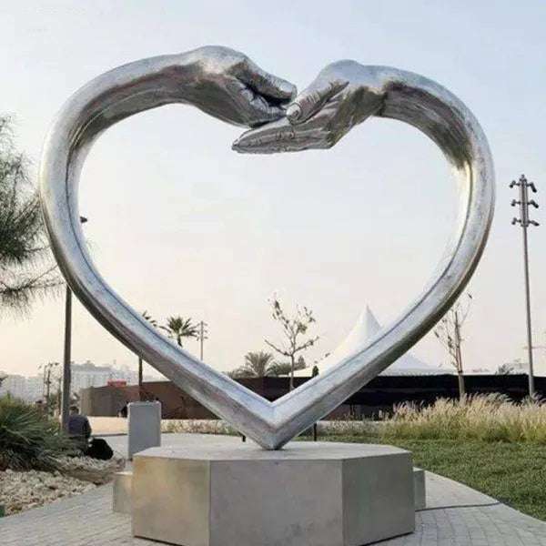 Symbolic metal heart artwork with raised hands touching at the top, set on a concrete base