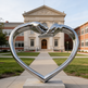 Metallic heart structure with sculpted hands meeting at the top, positioned in a landscaped courtyard