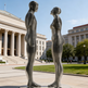Two metallic sculptures of human figures in front of a building with columns