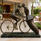 Bronze sculpture of a couple on a bicycle in an outdoor setting with greenery and a building interior.
Stylized bronze figures sharing a kiss beside a detailed bicycle