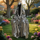 A ghost-like figure in a reflective silver hooded garment standing in a garden with flowers and trees.
Bronze artwork featuring a hooded figure with deep folds and shadows