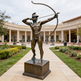 Bronze statue of a archer in a courtyard with a building in the background
Metal statue of a helmeted archer preparing to shoot an arrow