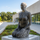 Modern sculpture of a seated figure in an outdoor setting with a building and trees in the background.