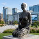 Modern sculpture of a woman in an urban setting with buildings and trees in the background