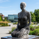 Modern sculpture of a seated figure in an outdoor setting with buildings and trees in the background