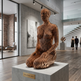 Wooden sculpture on a pedestal in an indoor setting with people and artwork in the background