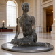Metallic sculpture of a seated figure in a grand hall with high ceilings and large windows.
