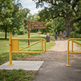 A yellow metal gate barrier stands at the entrance to a paved trail in Riverside Park, with a wooden signpost reading 'RIVERSIDE PARK TRAIL & GATE ACCESS' and two people walking in the background among trees.