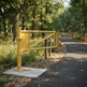 A yellow metal gate with a concrete base and a small sign, situated on a paved path in a wooded outdoor setting.