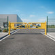 A yellow industrial automatic gate with green metal fencing on both sides, installed on a paved access road in front of a modern gray warehouse building under a blue sky.