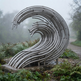 A large abstract metal wave sculpture made of curved silver tubes, mounted on a stone base in an outdoor garden or park with foggy trees and a path in the background.