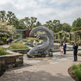 A large abstract wave-like metal sculpture stands in a landscaped garden courtyard with stone pathways, trees, a reflecting pool, and several people standing nearby. A bronze plaque is visible on the left.