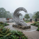 A large metallic wave-shaped sculpture stands on a stone base in a misty landscaped park with pathways, trees, and small visitors walking nearby.