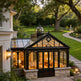 Orangery attached to a house with warm interior lighting, surrounded by greenery.