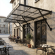 Outdoor cafe with tables and chairs under a large metal awning on a stone building.