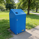 Blue trash bin labeled 'TAMCO' in a park setting with green grass and trees.
