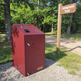 Red campground trash bin next to a wooden sign indicating 'Campground Trash' in a forested area.