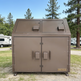 Brown metal trash bin with 'Taimco' branding in a park setting with trees and RVs.