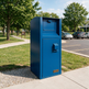 A tall blue metal mailbox or mail drop unit standing outdoors on a concrete sidewalk beside grass, trees, and a parking lot with cars.