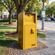 A bright yellow Larkoo outdoor mailbox with a top drawer and a lower door, situated on a paved sidewalk next to a grassy park area with trees and a street in the background.