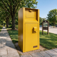 A bright yellow Taimco secure mailbox unit standing on a gravel bed beside a paved walkway, with trees, grass, and a parking lot sign in the background.