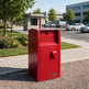 A red metal mailbox with a front door and top mail slot, placed outdoors on a gravel path beside a sidewalk and trees.