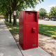 A red metal mailbox with a top slot and a lower door, standing on a concrete sidewalk outdoors near trees and grass.