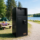 A black outdoor trash compactor unit with a front door and top opening, standing on a gravel path beside a lake with trees, a picnic table, and a tent in the background. A small bronze plate on the lower front reads TAIMCO.