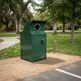 A green metal trash compactor bin with a circular opening at the top, set on a gravel path in a park with trees and grass in the background.