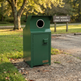 A dark green metal trash can with a circular opening at the top, shown outdoors on a gravel path near grass and trees. A small TAMACO label is visible on the front.