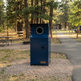 A dark blue metal trash compactor bin with a circular opening at the top, standing on a gravel path in a park with trees and picnic tables in the background.