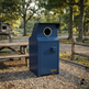 A dark blue metal trash compactor unit with a circular opening at the top, standing on a gravel surface in an outdoor park setting with picnic tables and trees in the background.