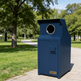 A dark blue metal trash can with a circular opening on top, standing on a paved path in a park with green trees and grass.