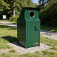 A dark green outdoor trash compactor with a circular opening on top and a small yellow TAIMCO brand plate on the lower right, situated on a gravel base in a grassy park area with a pathway and trees in the background.