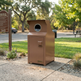 A brown metal recycling bin with a circular opening on top, placed on a concrete sidewalk in a park setting with trees, grass, and a recycling sign in the background.