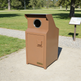A tall, brown metal trash can with a circular opening on top and a small front door, photographed outdoors on a paved path in a park. The brand name TAMCO is visible on a small badge near the bottom.