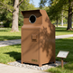 A large brown metal trash compactor unit with a circular opening on top, set outdoors on a gravel bed beside a paved path in a park. A small sign is placed nearby, and trees and grass are visible in the background.