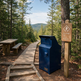 A dark blue public trash can stands beside a wooden trail sign reading 'Fine Ridge Trail Moderate' in a forest setting with a boardwalk path, picnic tables, and mountains in the background.