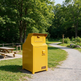 A bright yellow metal trash compactor unit standing on a gravel path in a park with trees, grass, picnic tables, and a small sign that appears to read TRACO.