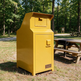 A bright yellow TALMO outdoor storage cabinet with a front door and top drawer, situated on grass in a wooded park area next to a picnic table.
