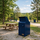 A dark blue metal mailbox or mail drop unit standing outdoors in a park-like setting with trees, a picnic table, lamp posts, and a gravel path.