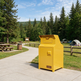 A bright yellow metal trash can with a hinged lid and front compartment, placed on a concrete path in a park with picnic tables, trees, and tents in the background.