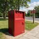 A red metal mailbox or mail collection cabinet standing outdoors on a gravel patch beside a sidewalk, with grass, trees, a parking lot, and a 'Public Garden - Keep Clean' sign in the background.