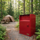 A red metal mailbox cabinet stands on a gravel campsite path in a wooded forest, with a tan tent and picnic table in the background.