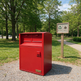 A red metal trash and recycling bin with two top compartments and a lower door, placed outdoors on a gravel path in a park next to a wooden sign post.